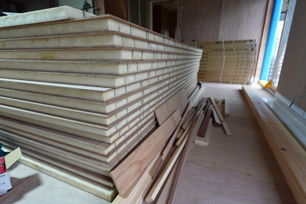 DSC03001 | みすずのリフォーム |四日市市の断熱リノベーション・性能向上リフォーム Stacks of drywall sheets with yellow padding and trim, piled in a partially finished room at a construction site with lumber on the floor.