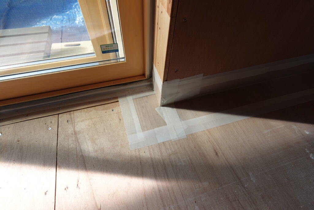 LDK窓付近床 | みすずのリフォーム |四日市市の断熱リノベーション・性能向上リフォーム Corner of a room under construction with taped floor protection along the baseboard and a wooden door frame beside a window stickered area; sun casts a shadow on the plywood floor.