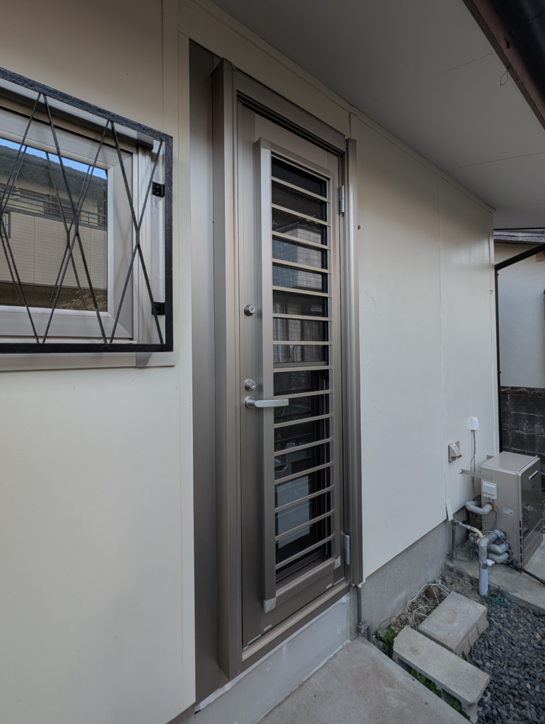 Exterior entry with a metal security door and a barred window on a light-colored wall; concrete steps and utilities nearby.
