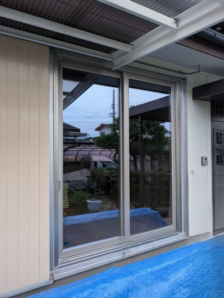 Sliding glass patio doors being installed, with a blue protective tarp on the ground and beige siding nearby.