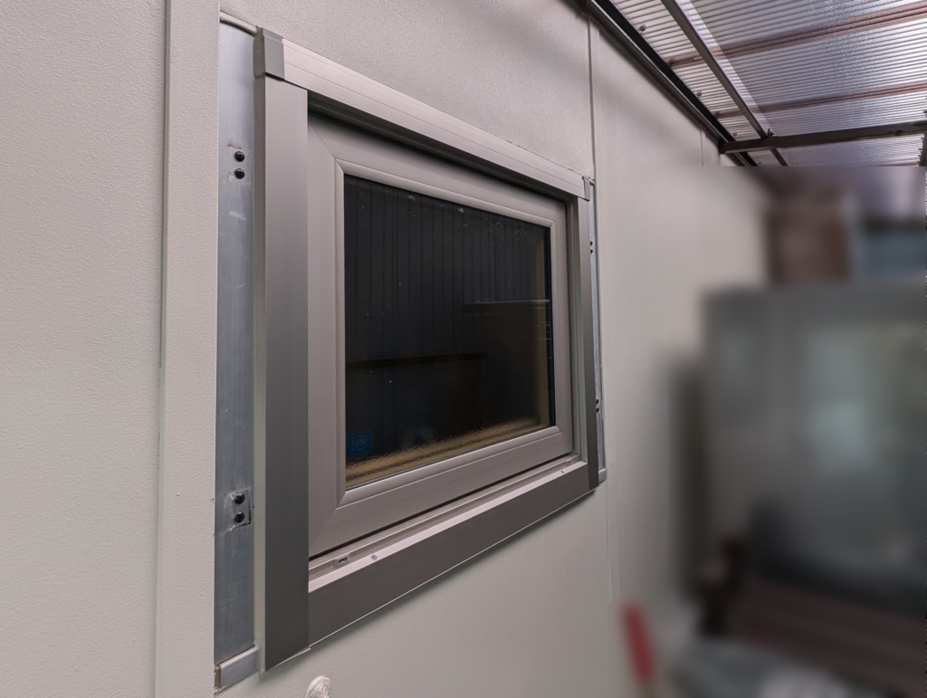 Window set in a light grey wall with a metallic frame and aluminum sash, under a translucent corrugated roof edge visible above.