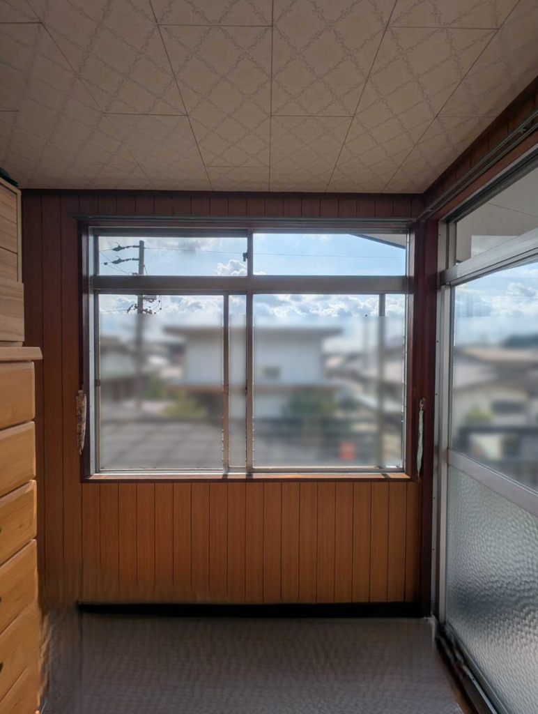 Sunlit room with wooden panel walls and a large window looking out at a suburban neighborhood.