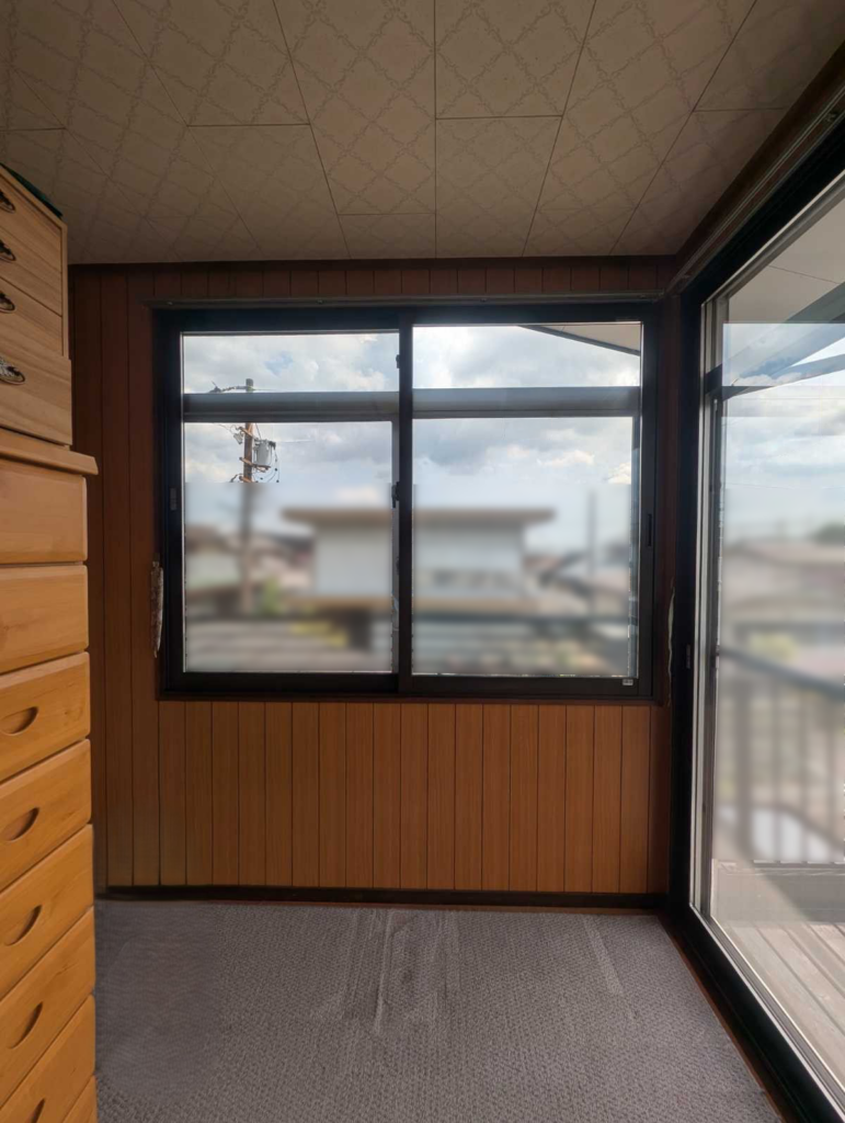 Interior of a small room with wooden panel walls, a tall dresser on the left, and a large window looking out to a cloudy sky and street scene outside.