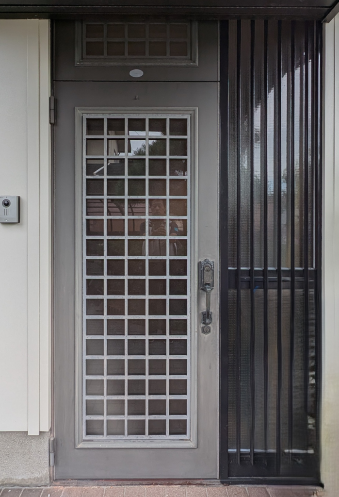 Gray exterior front door with a metal security grille featuring a square grid pattern and a separate glass door with vertical bars to the right.
